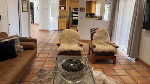 A living room with a sofa, two armchairs, a glass coffee table, and a view into the kitchen.