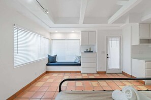 Quarto com cama, janela com persianas, um pequeno espaço de estar com almofadas, porta e cozinha compacta.