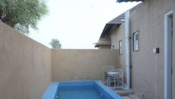 Una pequeña piscina exterior con borde de azulejos, una silla metálica y un edificio con tejado y puerta de azulejos.