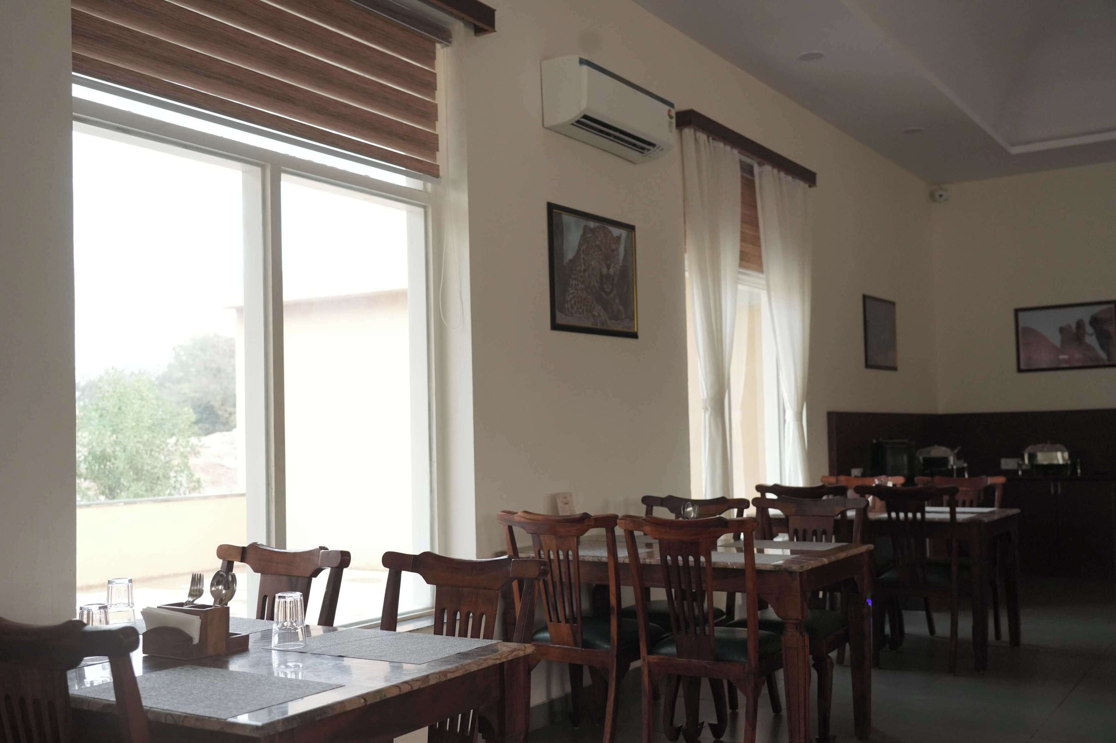 A dining area with wooden tables and chairs, large windows with blinds, and wall-mounted air conditioning units.