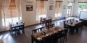 A dining area with multiple tables and chairs, large windows with curtains, and framed pictures on the walls.