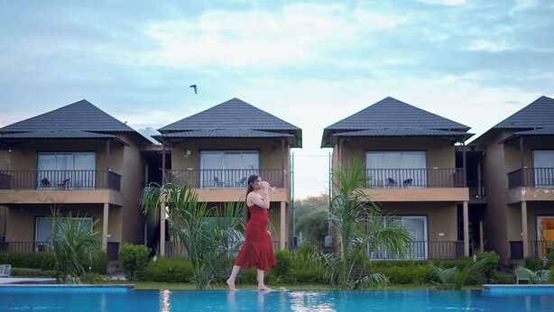 Une femme vêtue d’une robe rouge se tient près d’une piscine, avec une rangée d’immeubles en arrière-plan.
