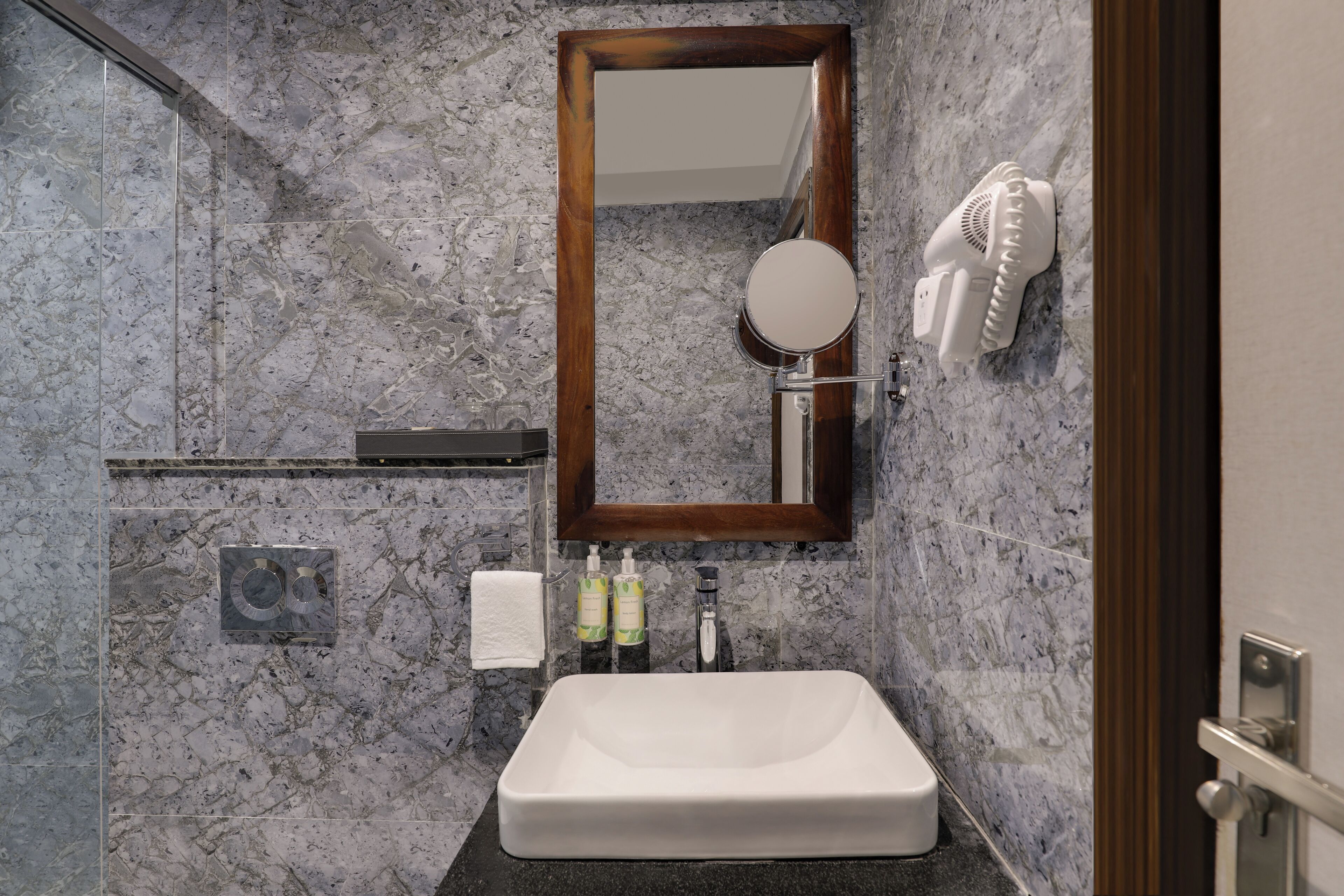 Un baño con pared de mármol, un espejo con marco de madera, un lavabo blanco, un perchero y un secador de pelo montado en la pared.