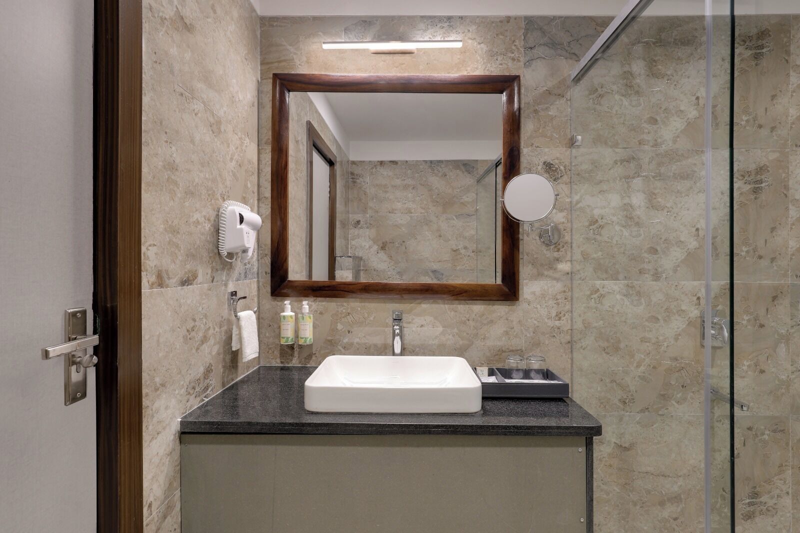 A bathroom with a marble wall, a large mirror, a sink, a hairdryer, and a shower.