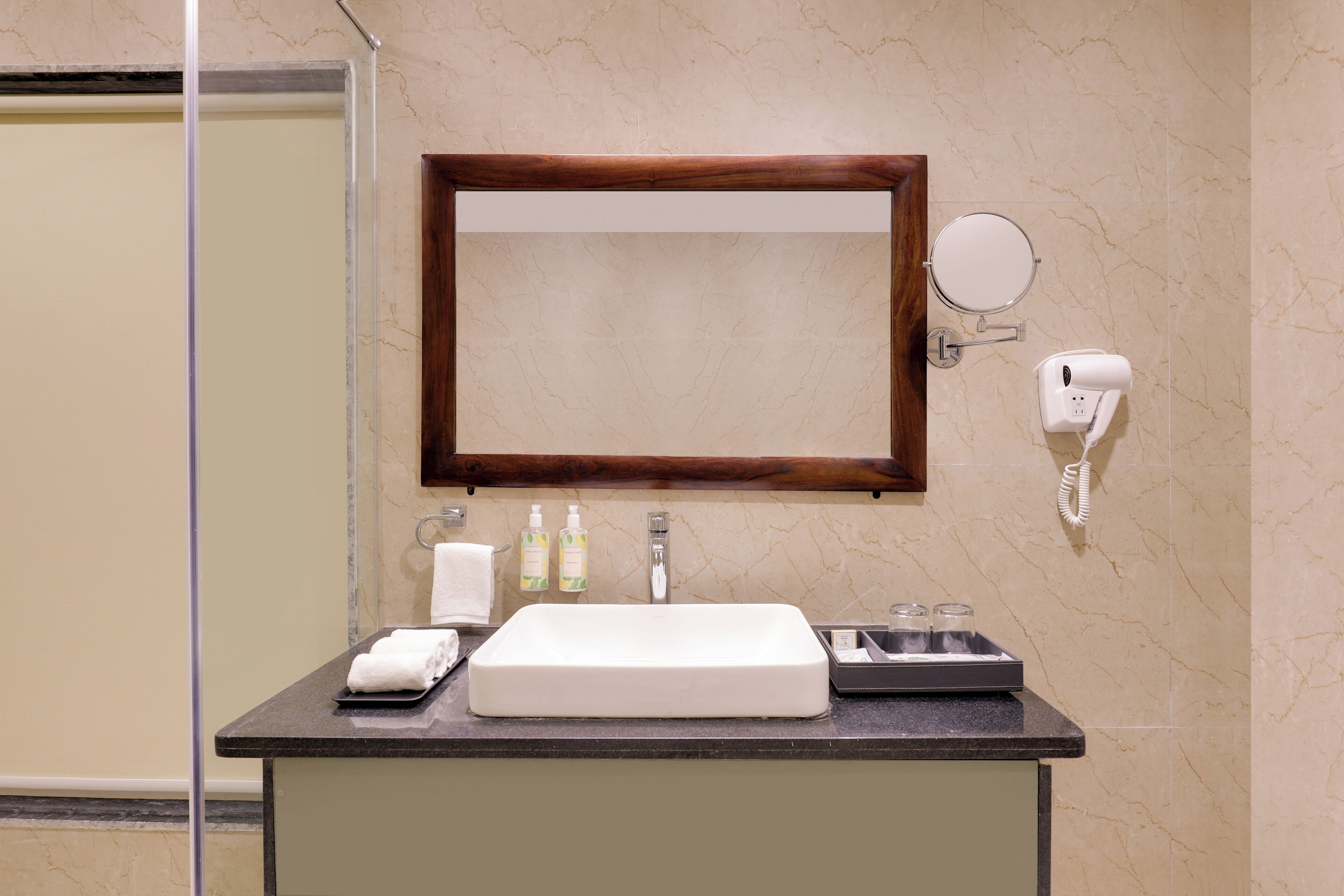A bathroom with a marble wall, a wooden-framed mirror, a white sink, a towel rack, a soap dispenser, and a hand dryer.