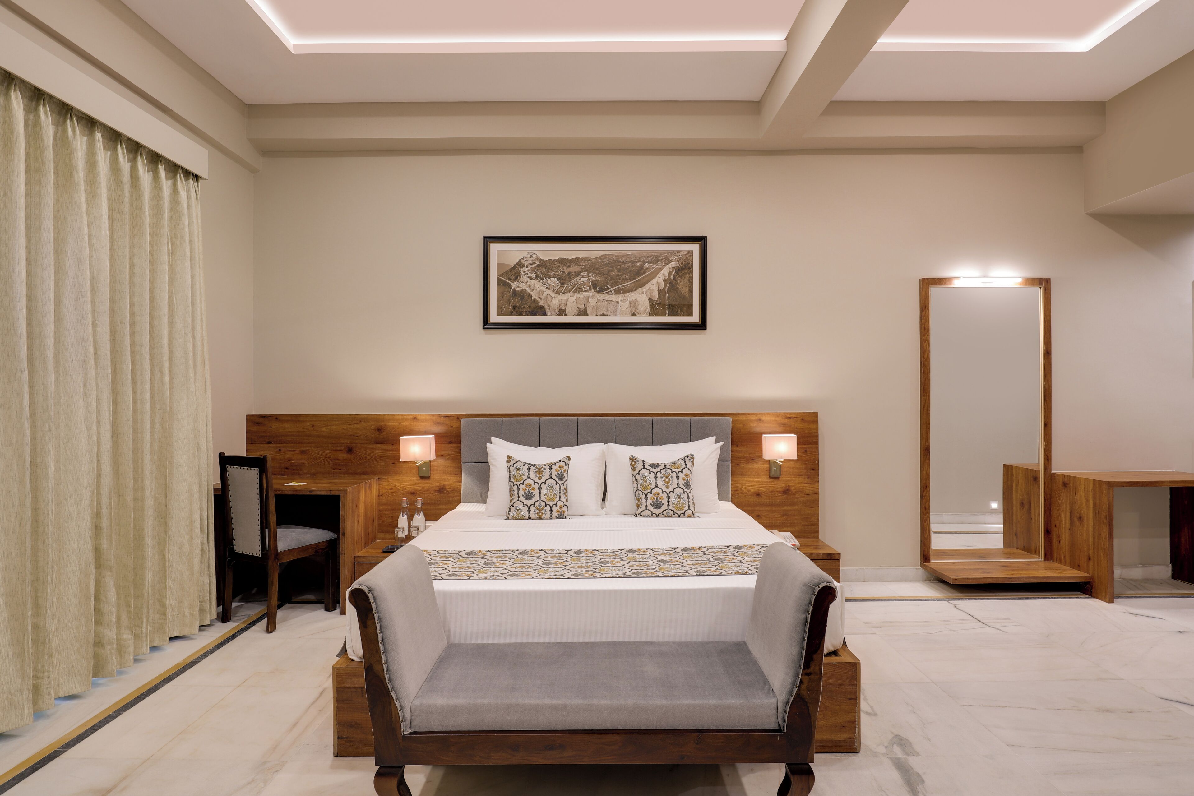 A modern hotel room with a large bed, a wooden headboard, a bedside table with a lamp, a large mirror, and a framed picture on the wall.