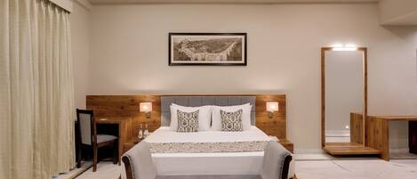 A modern hotel room with a large bed, a wooden headboard, a bedside table with a lamp, a large mirror, and a framed picture on the wall.