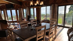 A dining area with a wooden table and chairs, large windows with a view of the ocean, and a hanging light fixture. - Magic Skies Main Level Living (Hilo)