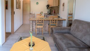 Un salon avec un canapé, un coin repas avec des chaises, une cuisine avec des meubles en bois et une horloge au mur.