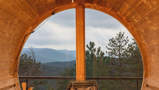 Une cabane en bois dotée d’une grande fenêtre circulaire qui offre une vue sur les montagnes et les arbres.