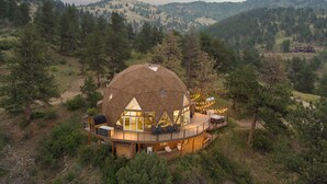 Un bâtiment en forme de dôme, doté d’une terrasse en bois et d’une grande fenêtre, entouré d’arbres et de collines.