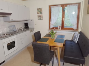 Een compacte keuken met witte kastjes, een koffiezetapparaat, en een eethoek met een tafel, stoelen en een bank.