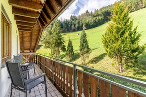 A balcony with a view of a green field and trees, wooden railing, and a table with chairs.