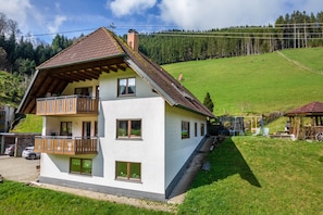 Een woonhuis met twee verdiepingen, een balkon, schoorsteen en uitzicht op een groene heuvel.