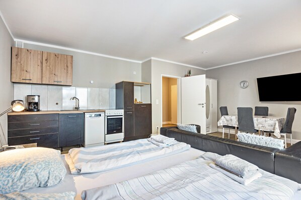 A modern bedroom with a large bed, a bedside table, a desk, a chair, a wardrobe, and a kitchenette.