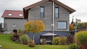 A two-story house with a grey exterior, a red-tiled roof, and a small front yard with a tree and a patio umbrella. - Holiday Apartment "Kathi‘s Allgäuliebe" with Mountain View, Private Terrace & Wi-Fi (Weitnau)