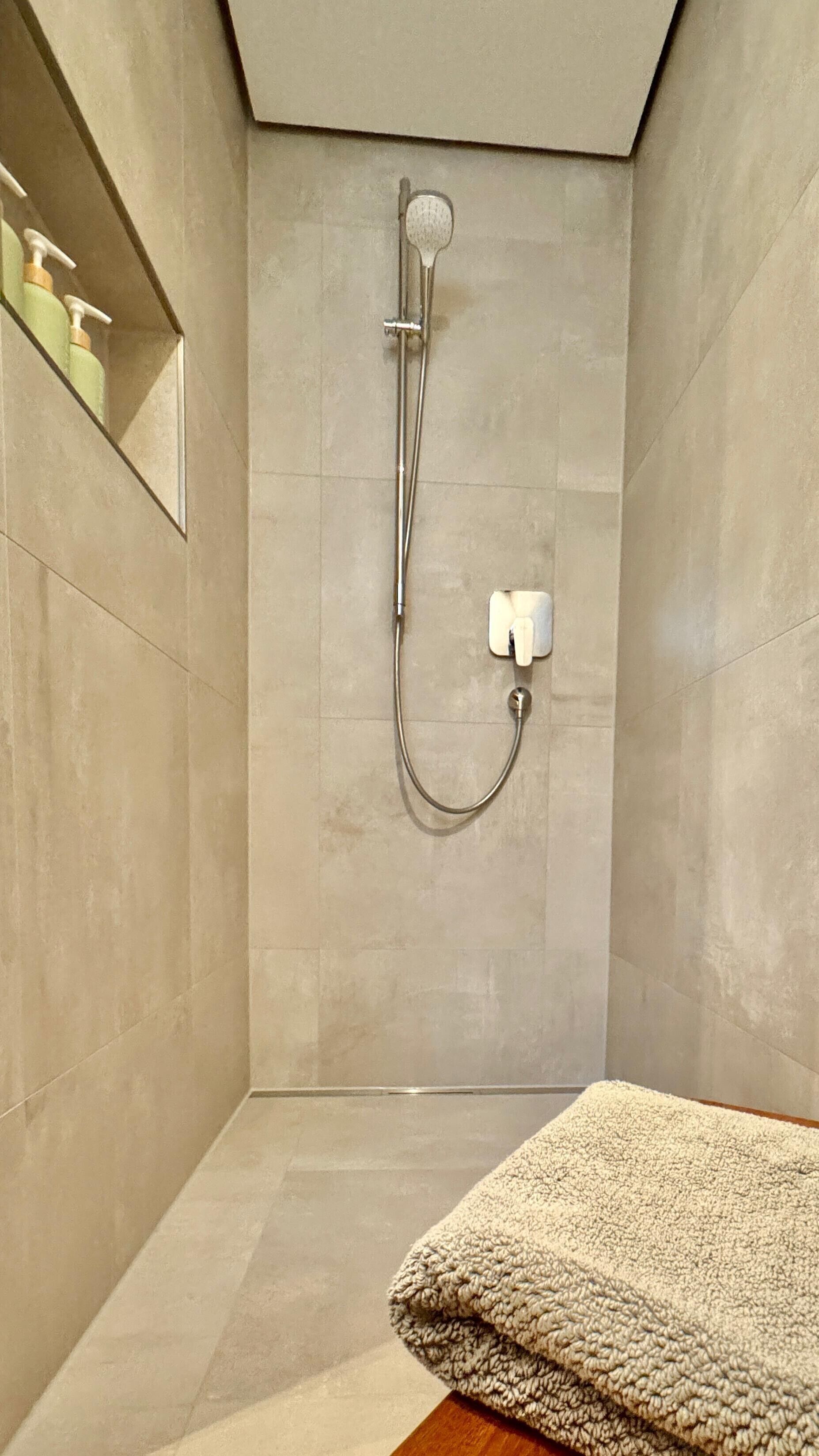 Eine Dusche mit Duschhandstück, Armaturen an der Wand und einem Handtuch auf einer Holzbank.
