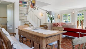Un coin repas comprenant une table en bois, des chaises, un banc, un escalier orné d’un tableau et une fenêtre avec des rideaux.
