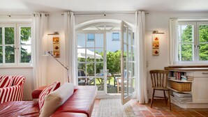 Une pièce avec un canapé rouge, une chaise en bois, un bureau avec des livres et une vue sur l’extérieur grâce à une porte vitrée.