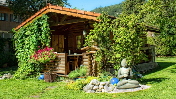 A rustic log cabin with a wooden porch, surrounded by lush greenery and a Buddha statue. - Holiday Apartment "Gästehaus Marianne" with Mountain View, Shared Terrace and Wi-Fi (Farchant)