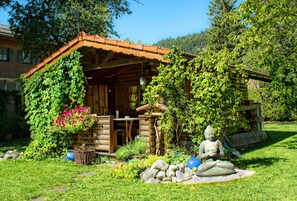 Une cabane rustique en rondins de bois, dotée d’un porche en bois, entourée d’une végétation luxuriante et ornée d’une statue de Bouddha.