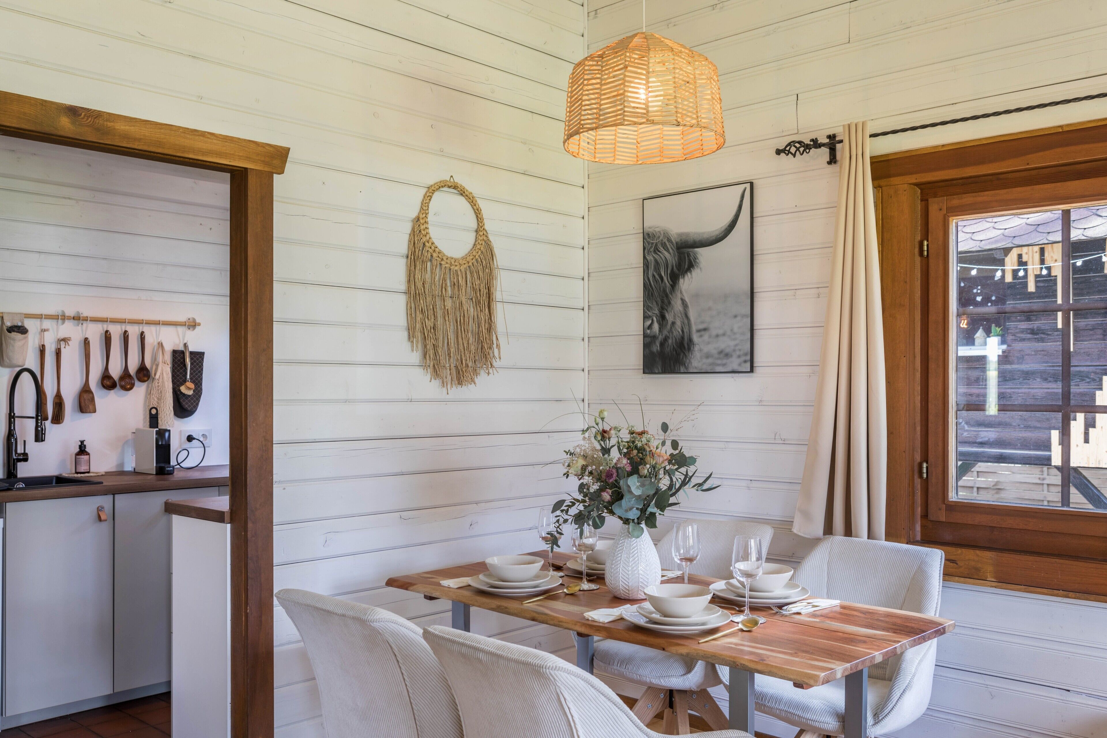 Un coin repas avec une table en bois dressée pour quatre personnes, des chaises blanches, un luminaire suspendu en fibres tressées, une décoration murale en macramé et un tableau encadré.
