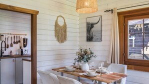 A dining area with a wooden table set for four, white chairs, a woven pendant light, a wall-mounted macrame piece, and a framed picture.