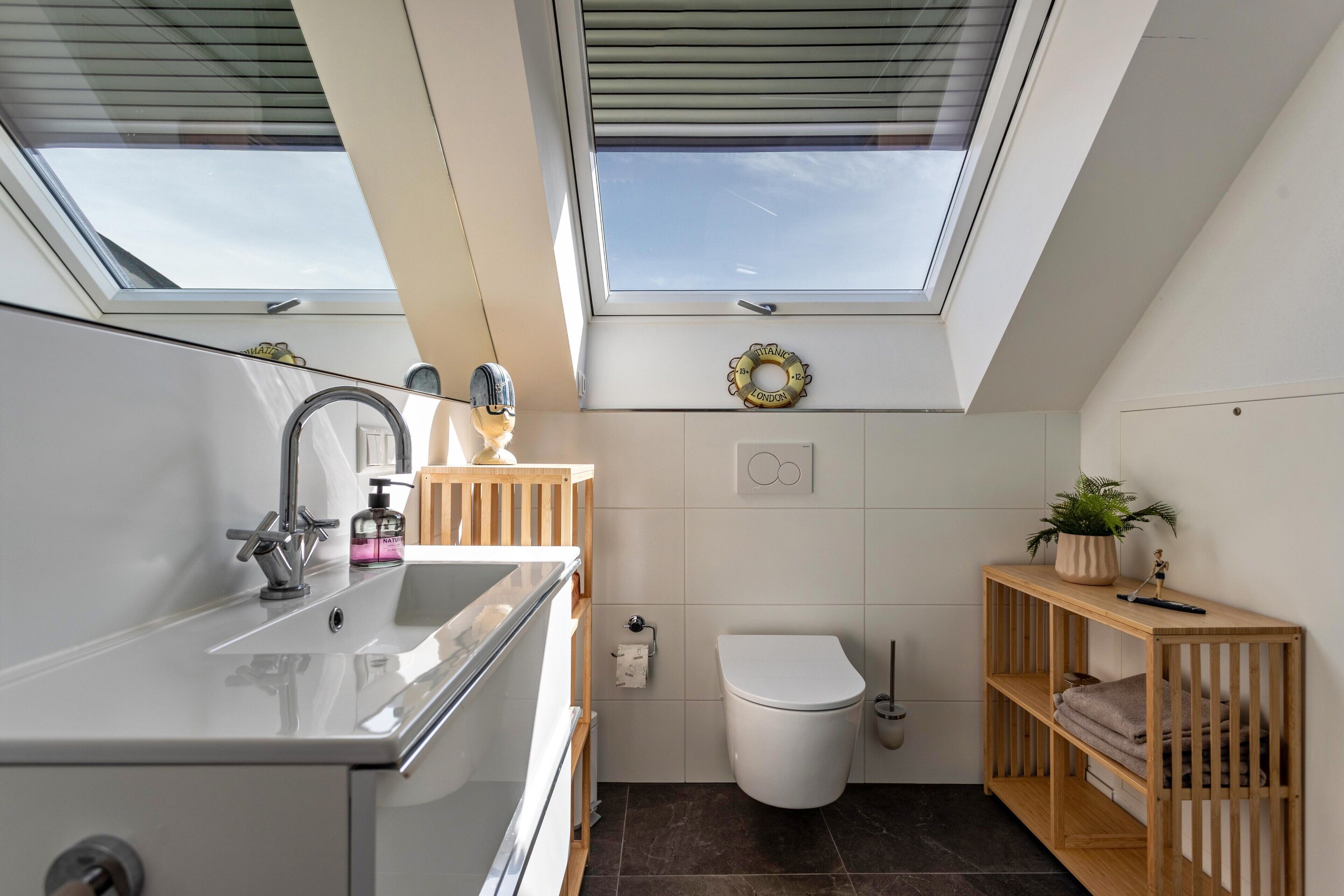 Ein modernes Badezimmer mit Dachfenstern, Toilette, Waschbecken und einem hölzernen Regal mit einer Pflanze.