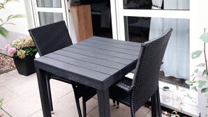 Une terrasse extérieure avec une table et des chaises, un pot de fleurs et une vue sur l’intérieur à travers une porte vitrée.