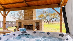 A hot tub under a wooden pergola with a stone fireplace in the background. - Hot tub, 290 Wine Trail, Covered Pavilion! (Fredericksburg)