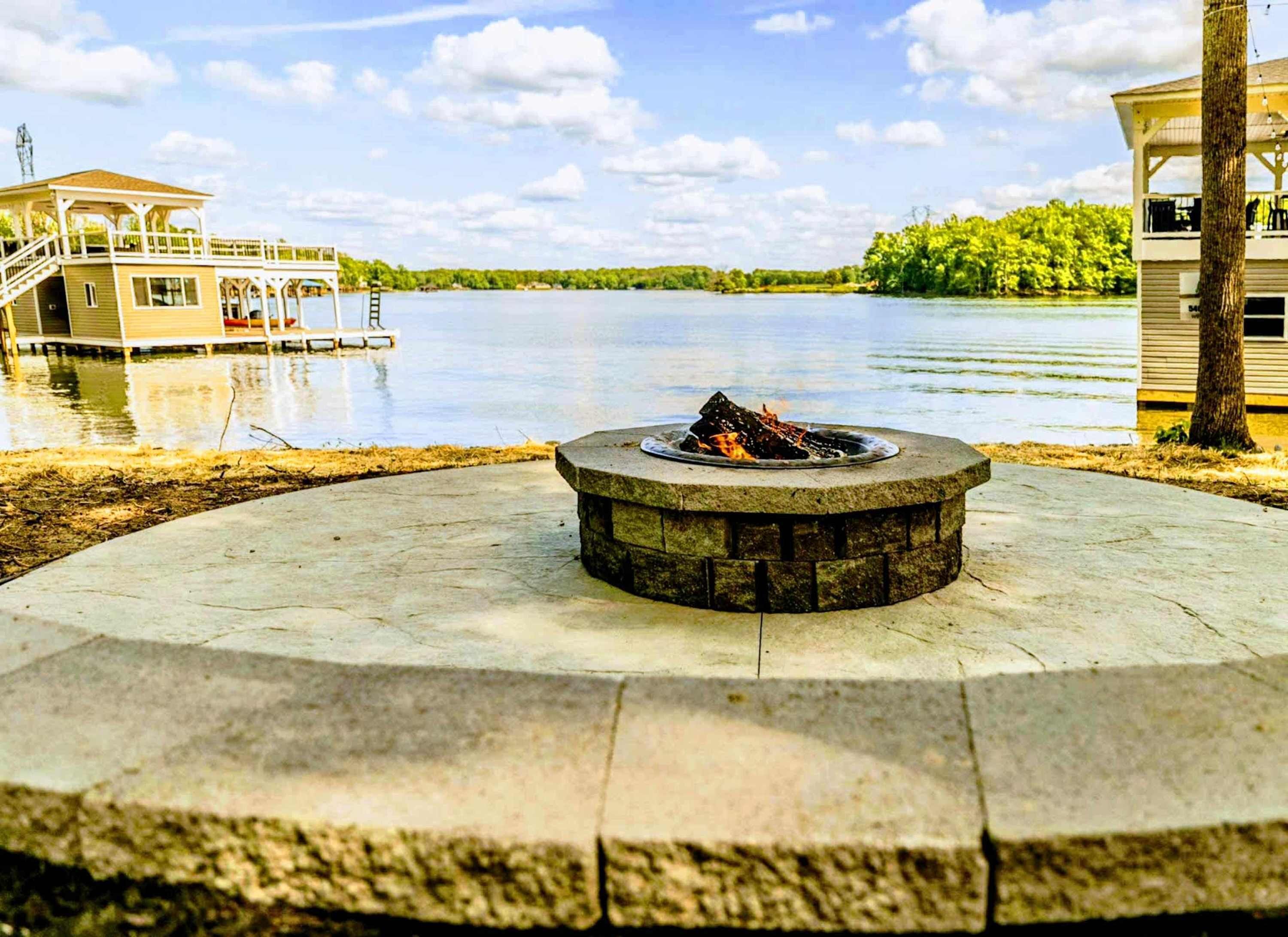 A circular stone fire pit with a fire, surrounded by a concrete deck, overlooking a lake with a dock and a building in the background.