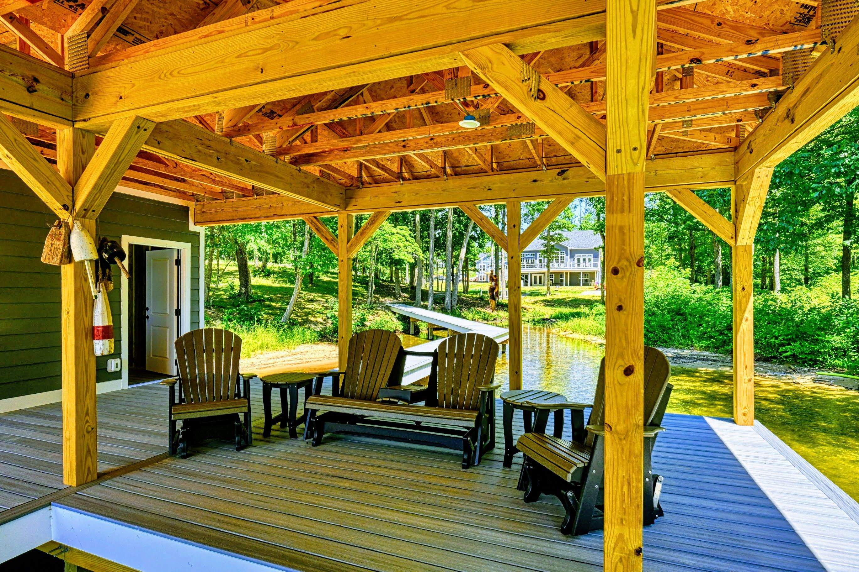A wooden deck with Adirondack chairs and a table, overlooking a body of water and a house.