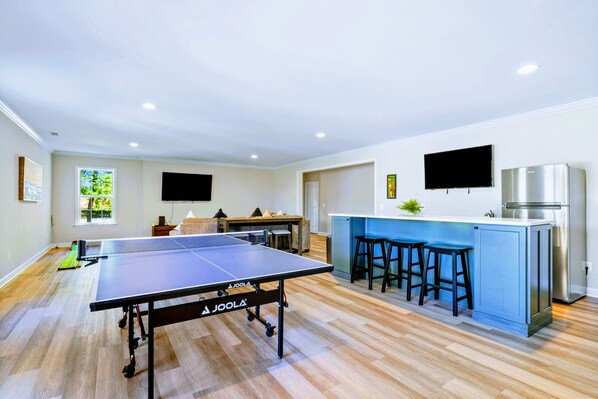A modern kitchen with a ping pong table, bar stools, a refrigerator, and a TV.