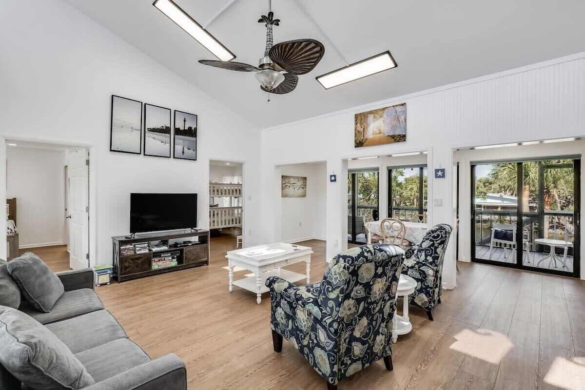 A spacious living room with a flat-screen TV, a ceiling fan, and a large window offering a view of the outdoors.