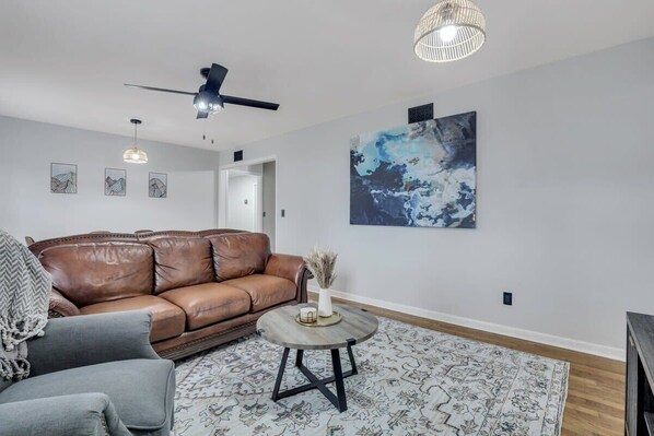 A living room with a brown leather sofa, a grey armchair, a round coffee table, and a patterned rug. There is a ceiling fan, pendant lights, and a large abstract painting on the wall. - Statesboro Sunshine Retreat (Statesboro)