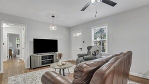 Een moderne woonkamer met een leren bank, een flatscreen televisie, een plafondventilator en een groot raam met jaloezieën.