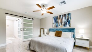 Un dormitorio con cama, ventilador de techo, mesita de noche, lámpara, un cuadro y una puerta corredera.