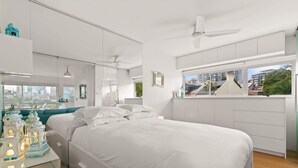 Un dormitorio moderno con una cama grande, un escritorio con objetos decorativos, vistas a la ciudad a través de la ventana y un ventilador de techo.