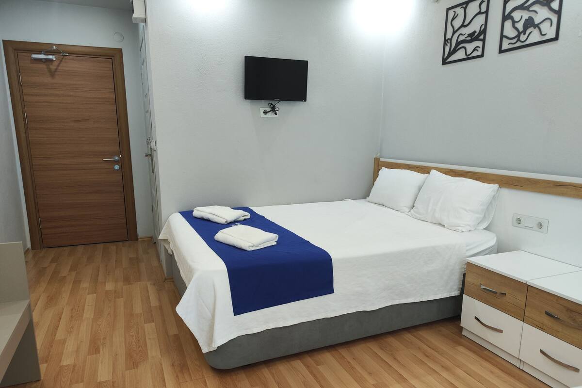 A neatly made bed with white and blue bedding, a wooden door, a bedside table with drawers, and a wall-mounted TV.