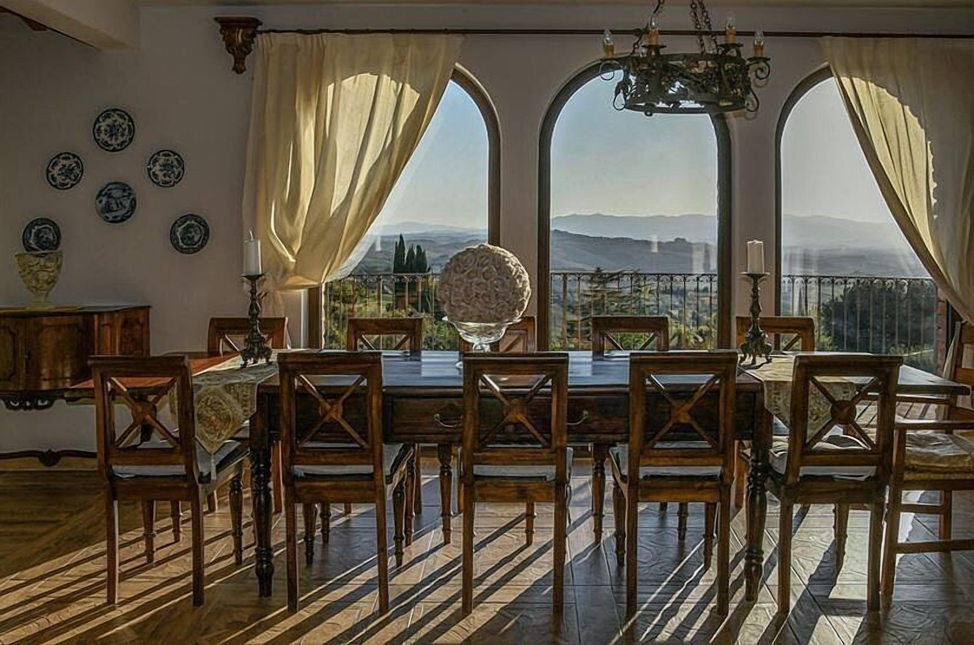 A dining room with a large table and chairs, a chandelier, and a view through arched windows.