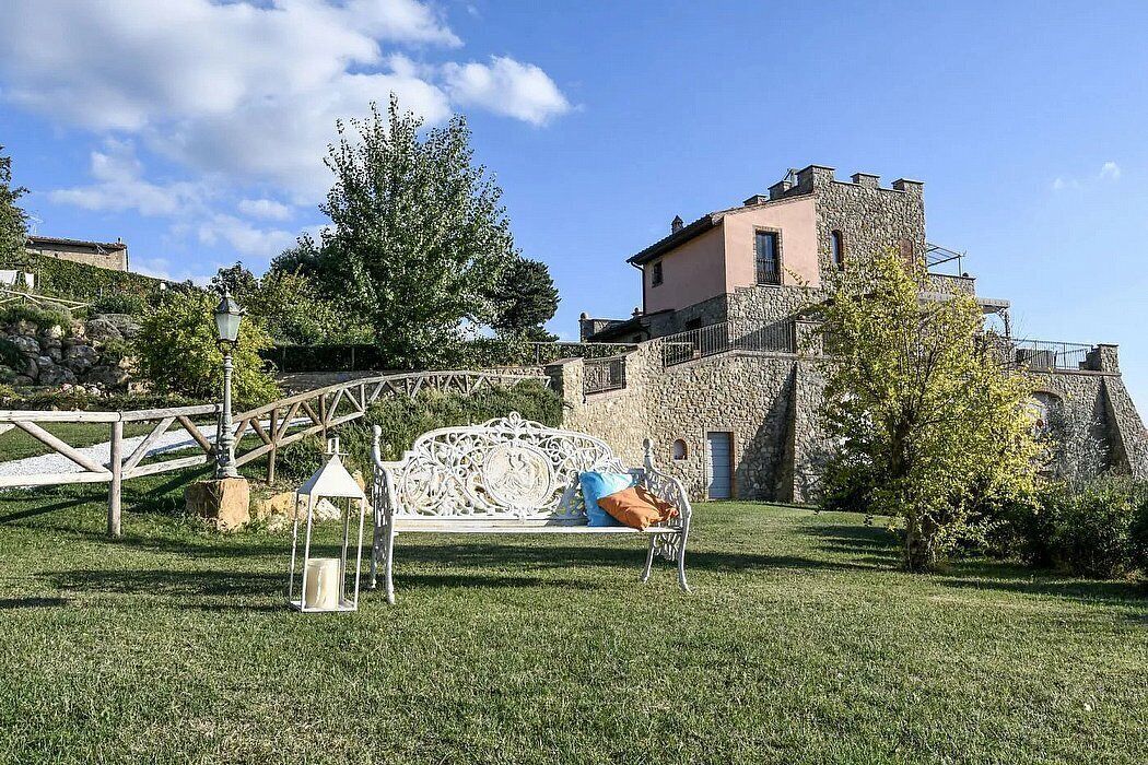 Un edificio in pietra con una torre, una panchina bianca con cuscini e un albero di fronte.