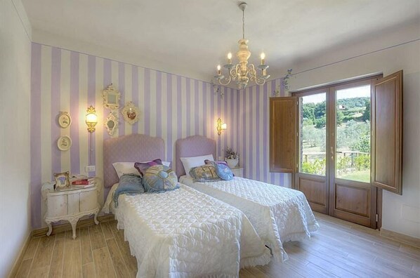 A bedroom with two beds, a chandelier, striped wallpaper, and a view of greenery through a large window.