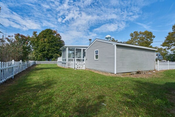A single-story mobile home with a fenced yard, a large tree, and a clear sky.