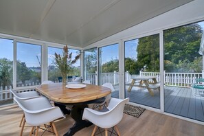 A covered patio with a wooden table and white chairs, large windows, and a view of a deck and trees.