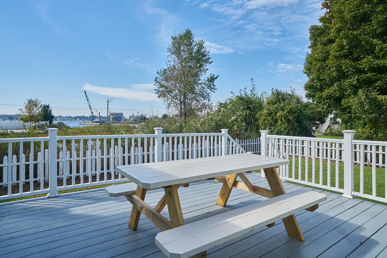 Eine Holzterrasse mit Picknicktisch, weißem Zaun und Blick auf Wasser und Bäume.