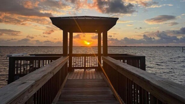 Un muelle de madera que conduce a una zona cubierta de observación, con un atardecer de fondo.