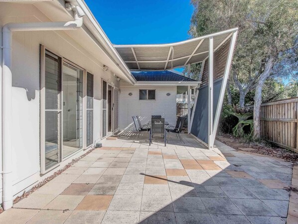 A covered patio area with a table and chairs, a sliding glass door, and a view of a backyard with a fence and trees. - 3 Bedroom Hastings Cove Villa! (Tweed)