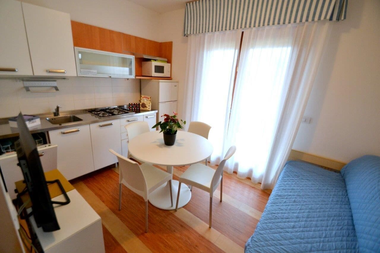 A compact kitchen with white cabinets, a microwave, and a stove. There's a dining table with chairs and a potted plant. A bed with a blue quilt is in the room.