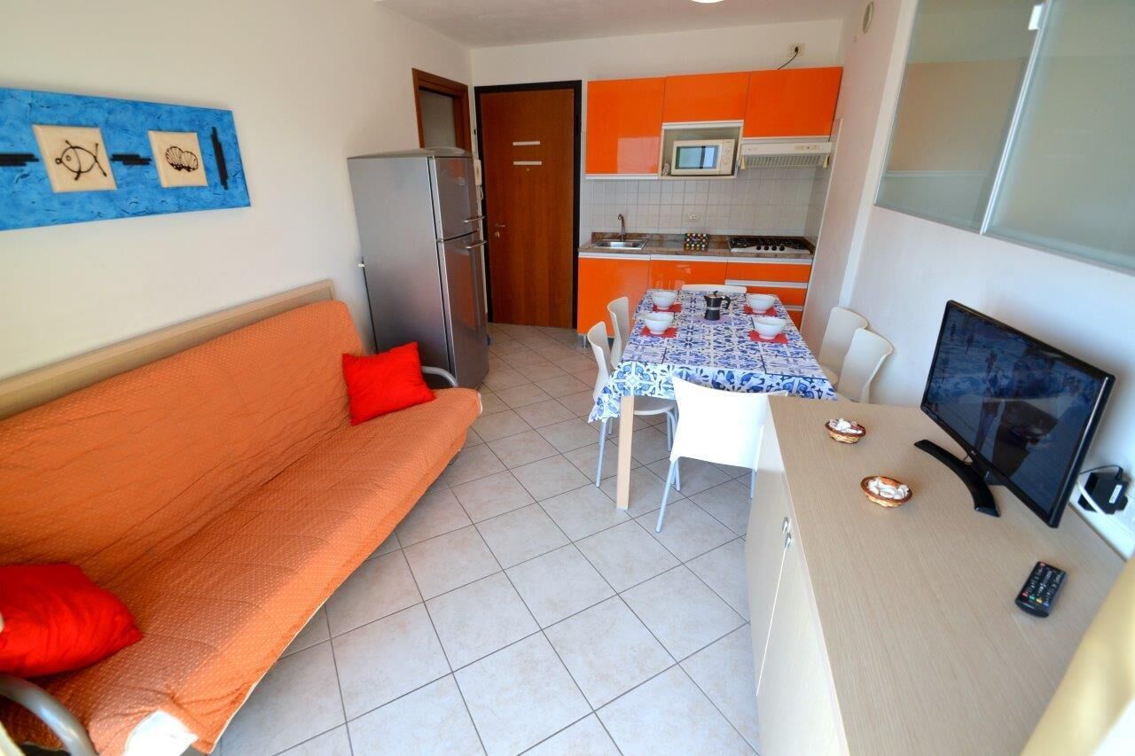 A compact living space with a sofa, a dining table with chairs, a kitchen area with orange cabinets, a refrigerator, and a television.