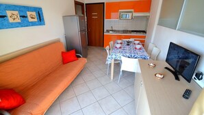 A compact living space with a sofa, a dining table with chairs, a kitchen area with orange cabinets, a refrigerator, and a television. - Luminous Flat 80m From the Beach - Beahost (San Michele al Tagliamento)